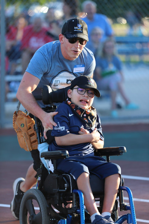 volunteer smiles and pushes boy in wheelchair across baseball field